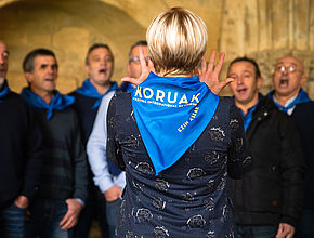 Une femme de dos avec un foulard bleu noué autour du cou, au centre de la photo, face à un chœur d'hommse. - Agrandir l'image (fenêtre modale)