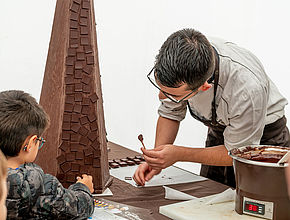 Un maître chocolatier à droite de laphoto derrière une table tend un batonnet avc une bouchée en chocolat à un petit garçon à gauche sur la photo. - Agrandir l'image (fenêtre modale)