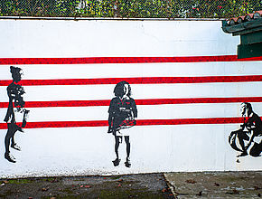 Sur un mur peint enblan trois personnages créés au pochoir avec une peinture noire, sont installés sur quatre lignes verticales rouges. - Irudia handitu (modu-leihoa)