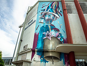 Une fresque murale représentant, dans les tons bleus, un homme coiffé d'un chapeau à lanières avec une cloche dans le dos. - Agrandir l'image (fenêtre modale)