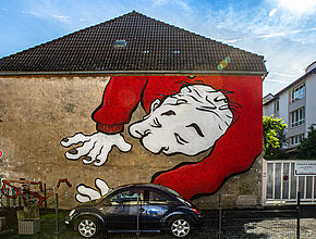 Au prmeier plan une voiture devant le mur d'une maison sur lequel, sur la droite est représenté un homme vêtu de rouge en train de chercher ses lunettes tombées dans le coin en bas à gauche du mur. - Irudia handitu (modu-leihoa)