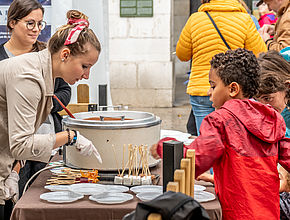A gauche, des jeuens filles munies de gants préparent des bouchées au chocolat prêtes à la dégustation sous l'oeil attentif des enfants, à droite de la photo. - Agrandir l'image (fenêtre modale)