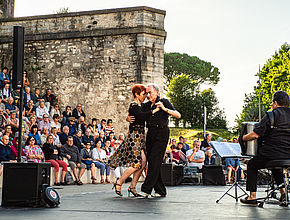 Un homme et une femme au centre de la photo font une démonstration de tango accompagnés par des musiciens sous le regard du public nombreux et assis sur la gauche. - Irudia handitu (modu-leihoa)